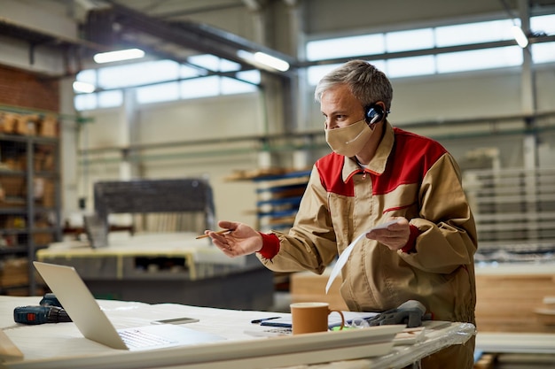 Mid adult carpenter with face mask having video call over laptop in a workshop