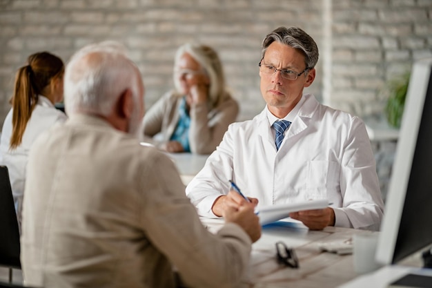 Mid adult doctor listening to his senior patient talk about health issues and writing notes in medical record at clinic