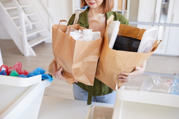 Mid section portrait of modern young woman holding paper bags with plastic items while sorting waste at home