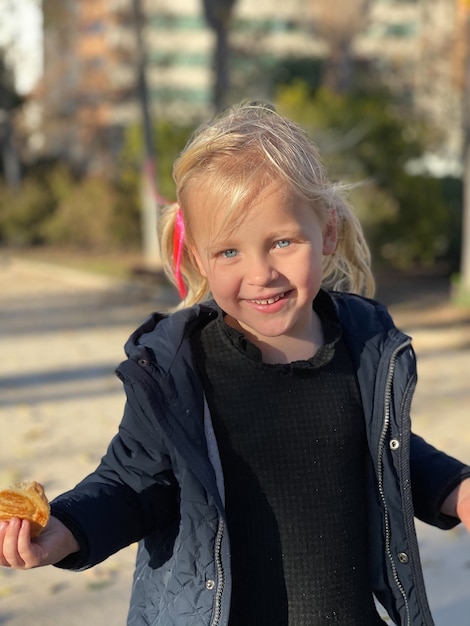 Mid shot of a cheerful toddler girl with a bun in hand. Outdoor portrait of blue-eyed child with fair hair.