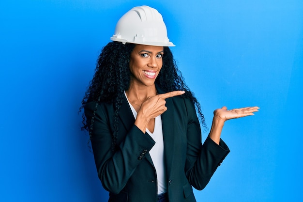 Middle age african american woman wearing architect hardhat amazed and smiling to the camera while presenting with hand and pointing with finger.