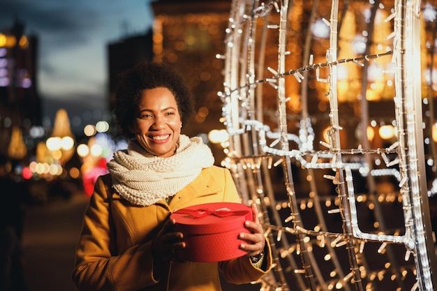 Middle age black woman holding red gift box while walking on the Christmas winter market in the city.