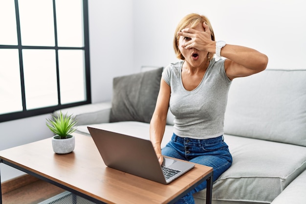 Middle age blonde woman using laptop at home peeking in shock covering face and eyes with hand looking through fingers with embarrassed expression