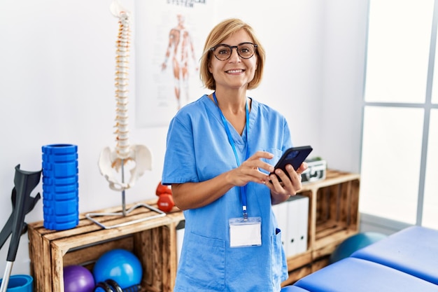 Middle age blonde woman wearing physiotherapist uniform using smartphone at clinic