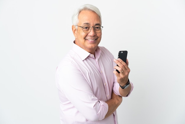 Middle age Brazilian man isolated on white background holding a mobile phone and with arms crossed