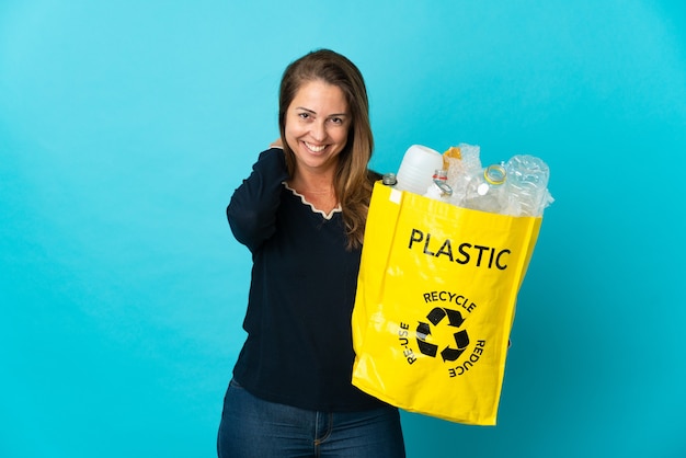 Middle age Brazilian woman holding a bag full of plastic bottles to recycle on blue laughing