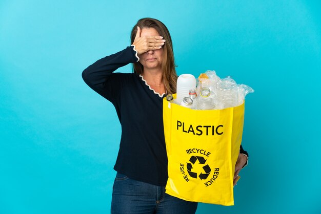 Middle age Brazilian woman holding a bag full of plastic bottles to recycle on blue wall covering eyes by hands. Do not want to see something