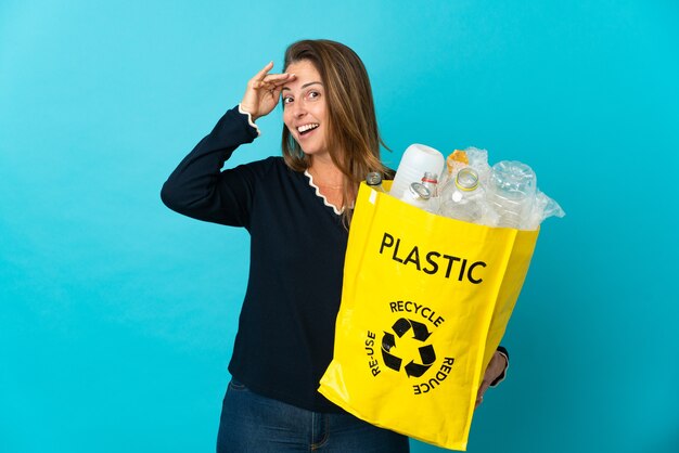 Middle age Brazilian woman holding a bag full of plastic bottles to recycle on blue wall doing surprise gesture while looking to the side