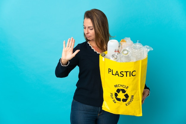 Middle age Brazilian woman holding a bag full of plastic bottles to recycle on blue wall making stop gesture and disappointed