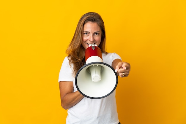 Middle age brazilian woman isolated on yellow shouting through a megaphone to announce something while pointing to the front