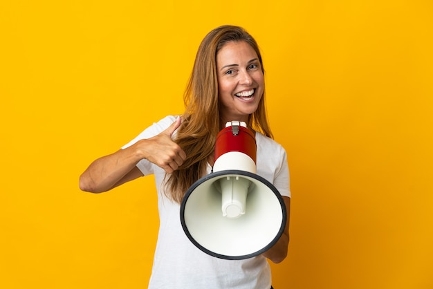 Middle age brazilian woman isolated on yellow wall holding a megaphone with thumb up