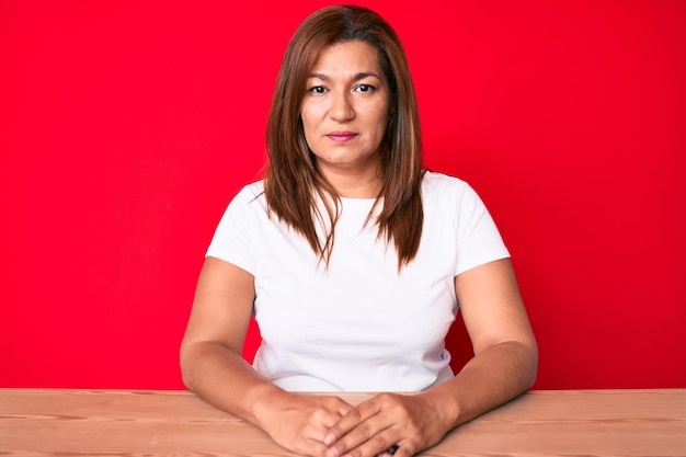 Middle age brunette hispanic woman wearing casual style sitting on the table with serious expression on face. simple and natural looking at the camera.