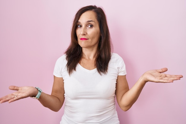 Middle age brunette woman standing over pink background clueless and confused with open arms no idea concept