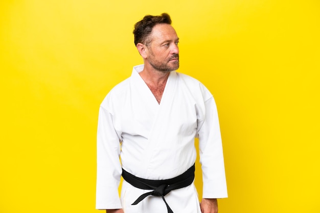 Middle age caucasian man doing karate isolated on yellow background looking to the side