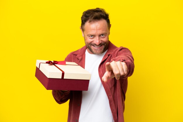 Middle age caucasian man holding a gift isolated on yellow background points finger at you with a confident expression