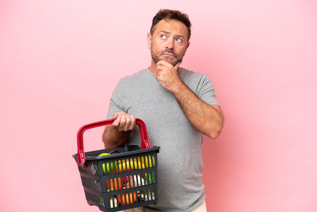 Middle age caucasian man holding a shopping basket isolated on pink background having doubts