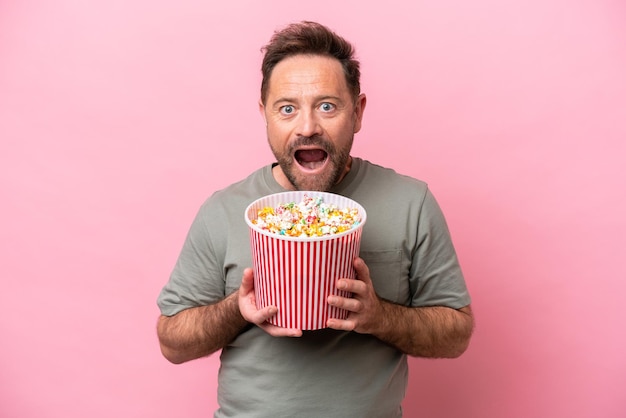 Middle age caucasian man isolated on pink background holding a big bucket of popcorns