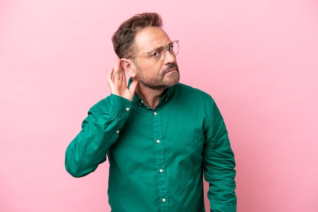 Middle age caucasian man isolated on pink background listening to something by putting hand on the ear