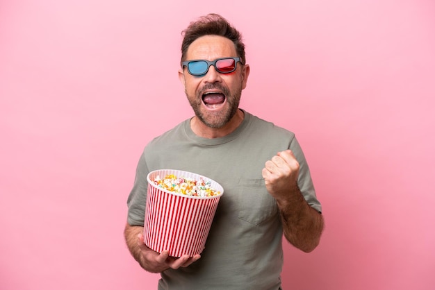 Middle age caucasian man isolated on pink background with 3d glasses and holding a big bucket of popcorns