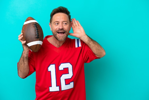 Middle age caucasian man playing rugby isolated on blue background listening to something by putting hand on the ear