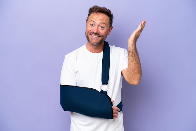 Middle age caucasian man with broken arm and wearing a sling isolated on purple background extending hands to the side for inviting to come