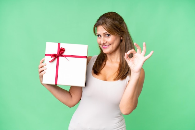 Middle age caucasian woman holding a gift over isolated background showing ok sign with fingers