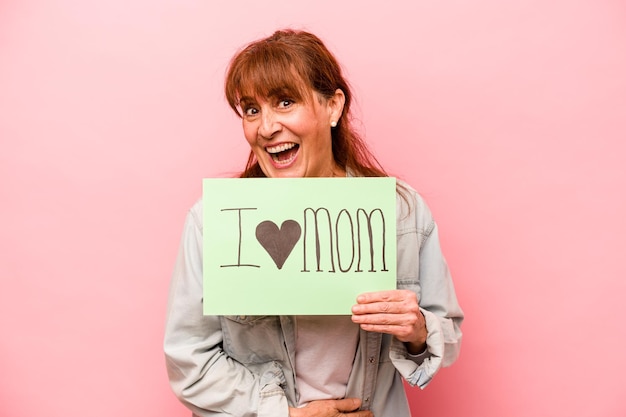 Middle age caucasian woman holding I love mom placard isolated on pink background laughing and having fun