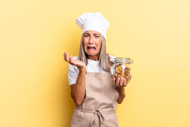 Middle age chef woman looking desperate and frustrated, stressed, unhappy and annoyed, shouting and screaming with cookies