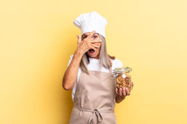 Middle age chef woman looking shocked, scared or terrified, covering face with hand and peeking between fingers with cookies