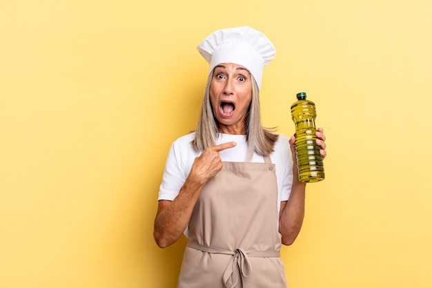 Middle age chef woman looking shocked and surprised with mouth wide open, pointing to self