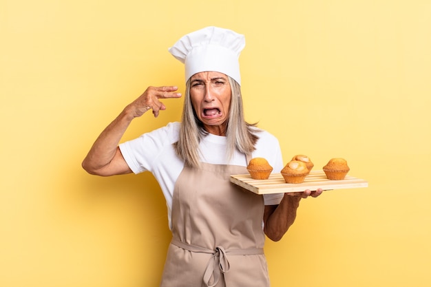 Middle age chef woman looking unhappy and stressed, suicide gesture making gun sign with hand, pointing to head and holding a muffins tray