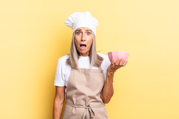 Middle age chef woman looking very shocked or surprised, staring with open mouth saying wow and holding an empty pot