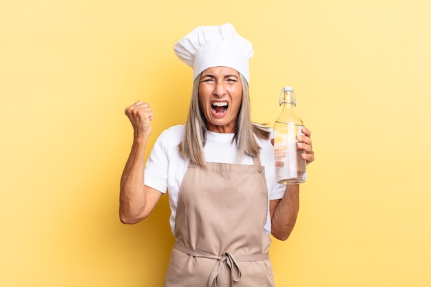 Middle age chef woman shouting aggressively with an angry expression or with fists clenched celebrating success with a water bottle