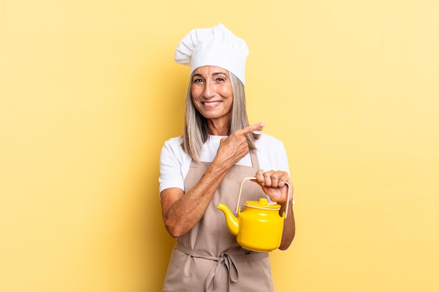 Middle age chef woman smiling cheerfully, feeling happy and pointing to the side and upwards, showing object in copy space and holding a teapot