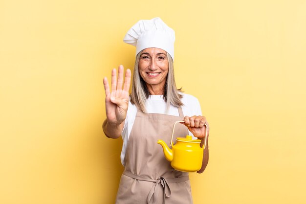 Photo middle age chef woman smiling and looking friendly, showing number four or fourth with hand forward, counting down and holding a teapot