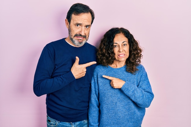 Middle age couple of hispanic woman and man hugging and standing together pointing aside worried and nervous with forefinger, concerned and surprised expression