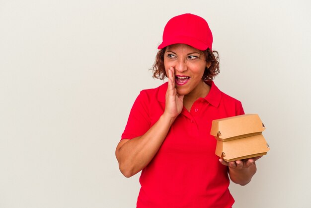 Middle age delivery woman taking burguers isolated on white background is saying a secret hot braking news and looking aside