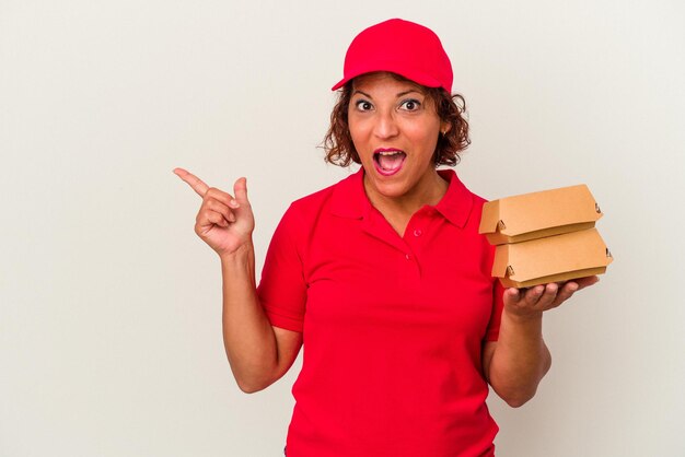 Middle age delivery woman taking burguers isolated on white background pointing to the side