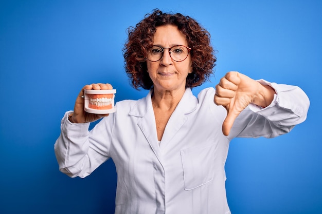 Middle age dentist woman wearing coat holding plastic denture teeth over blue background with angry face negative sign showing dislike with thumbs down rejection concept