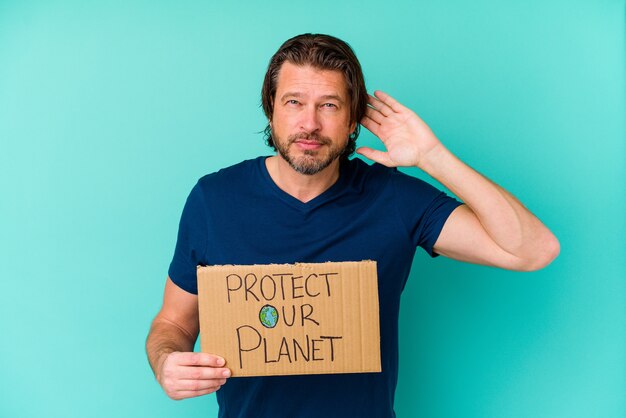 Middle age dutch man holding a protect our planet placard isolated on blue background trying to listening a gossip.