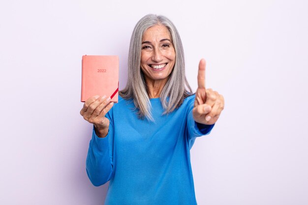 Middle age gray hair woman smiling proudly and confidently making number one. 2022 agenda concept