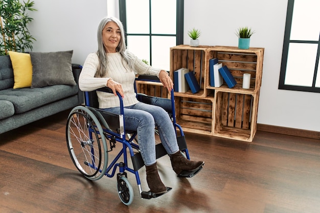 Middle age grey-haired woman smiling confident sitting on wheelchair at home