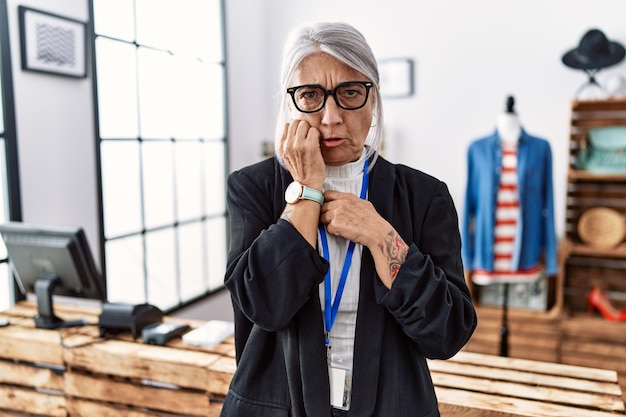 Middle age grey-haired woman working as manager at retail boutique looking stressed and nervous with hands on mouth biting nails. anxiety problem.