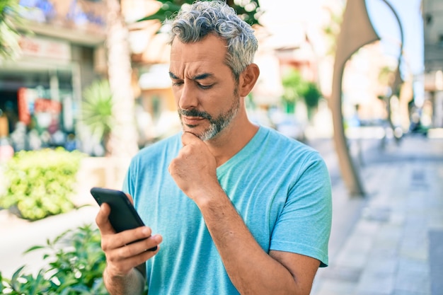 Middle age greyhaired man using smartphone at street of city serious face thinking about question with hand on chin thoughtful about confusing idea