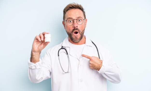 Middle age handsome man looking shocked and surprised with mouth wide open, pointing to self. physician with pills bottle