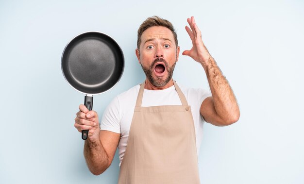 Middle age handsome man screaming with hands up in the air. frying pan concept