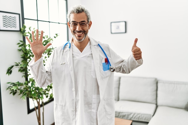 Middle age hispanic man wearing doctor uniform and stethoscope at waiting room showing and pointing up with fingers number six while smiling confident and happy.