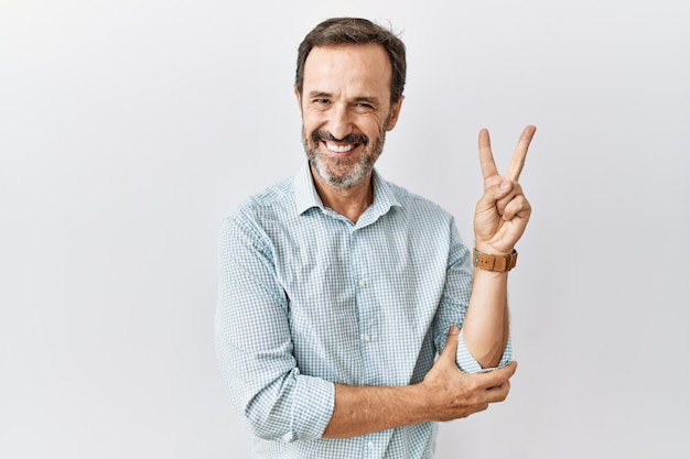 Middle age hispanic man with beard standing over isolated background smiling with happy face winking at the camera doing victory sign with fingers number two