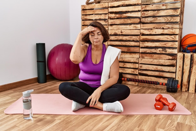 Middle age hispanic woman sitting on training mat at the gym worried and stressed about a problem with hand on forehead nervous and anxious for crisis