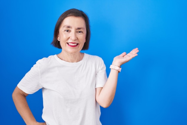 Middle age hispanic woman standing over blue background smiling cheerful presenting and pointing with palm of hand looking at the camera.